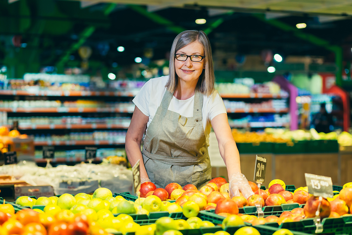 Portrait of senior woman in glasses at grocery store employee, supermarket. Stands in work clothes, near fruits, looks at the camera, smiles