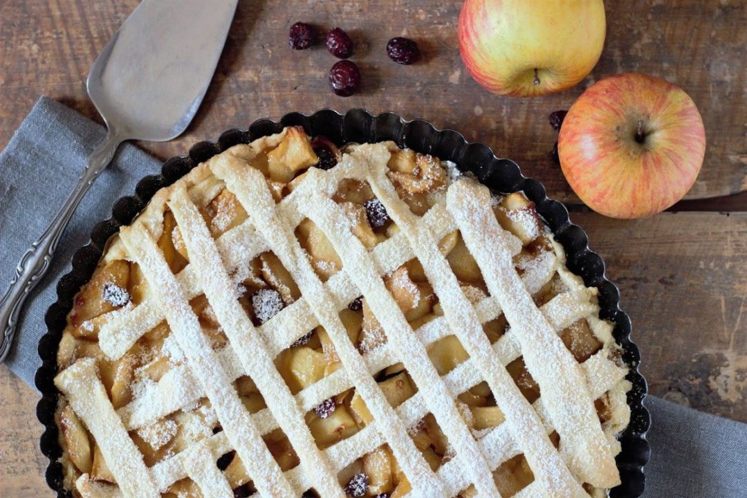 apple pie on wooden surface next to serving spatula and two apples