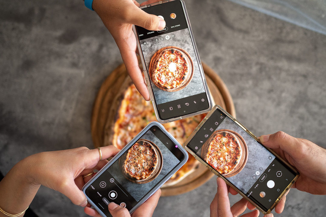 three hands holding phones and taking photos of pizza