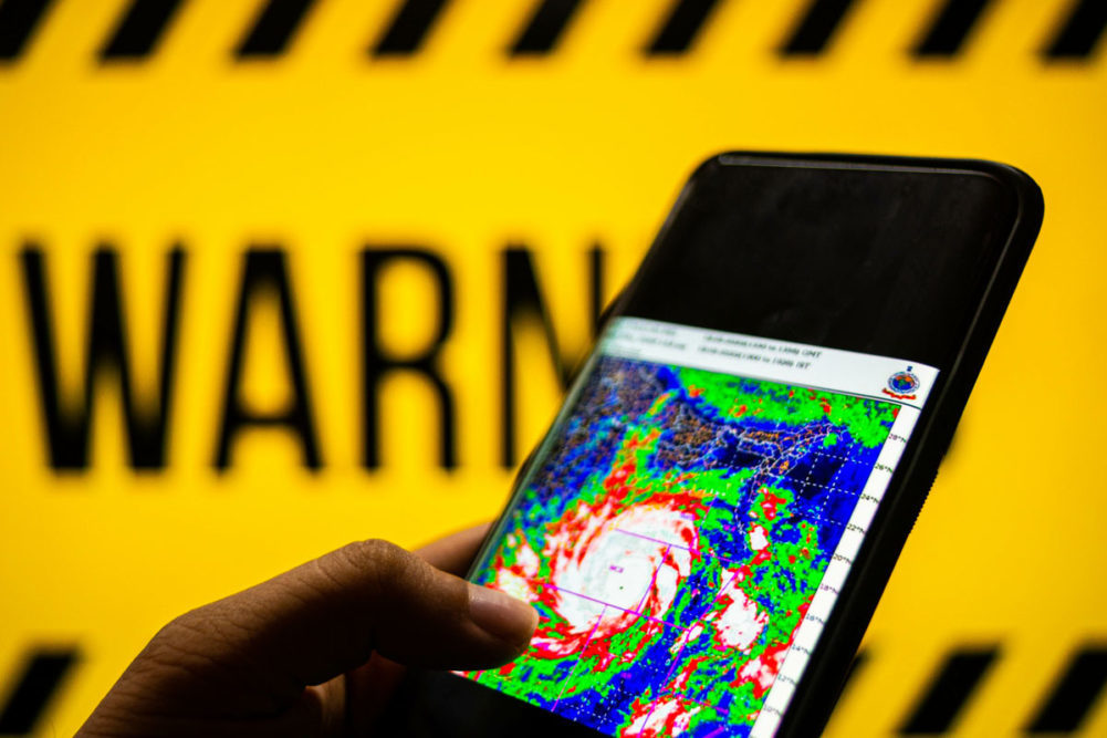 hand holding a smart phone with a hurricane map on the screen and a warning sign in the background