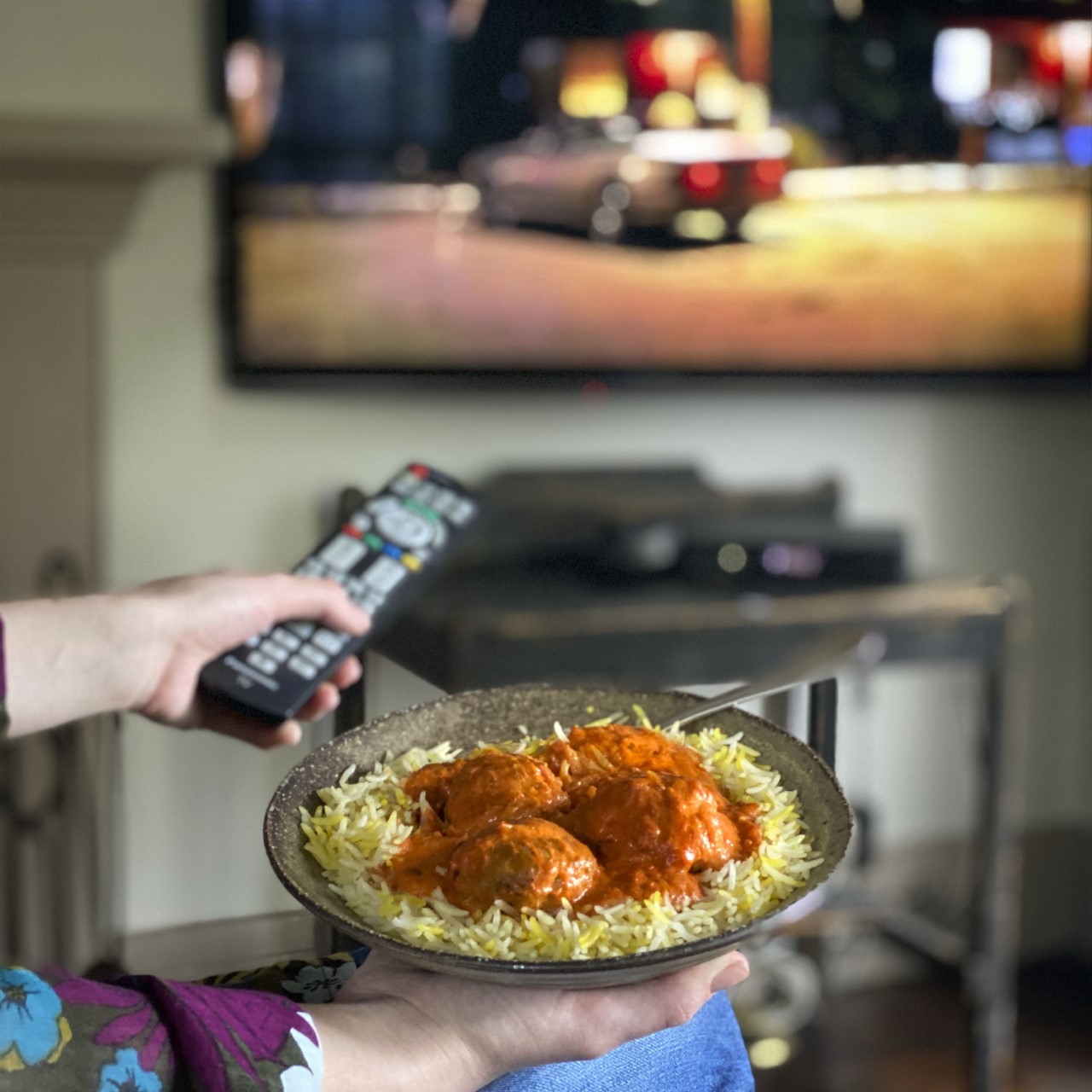someone holding a plate with an Indian meal on it in one hand and a TV remote in the other hand, pointing it at a TV on a wall