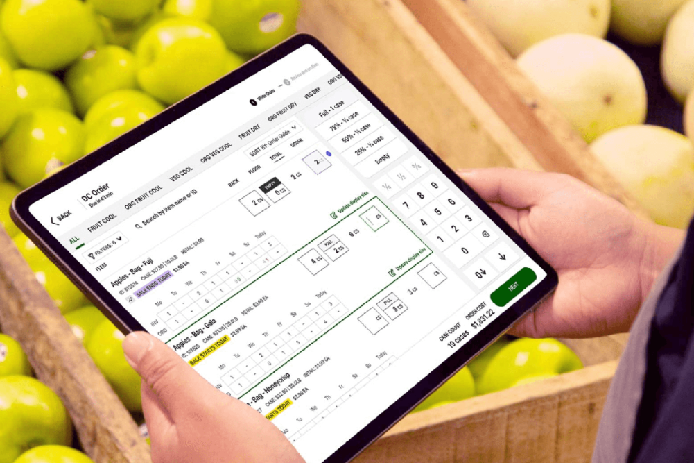 hands holding a tablet with supply chain technology on the screen. The tablet is being held in front of a produce stand with fruit.