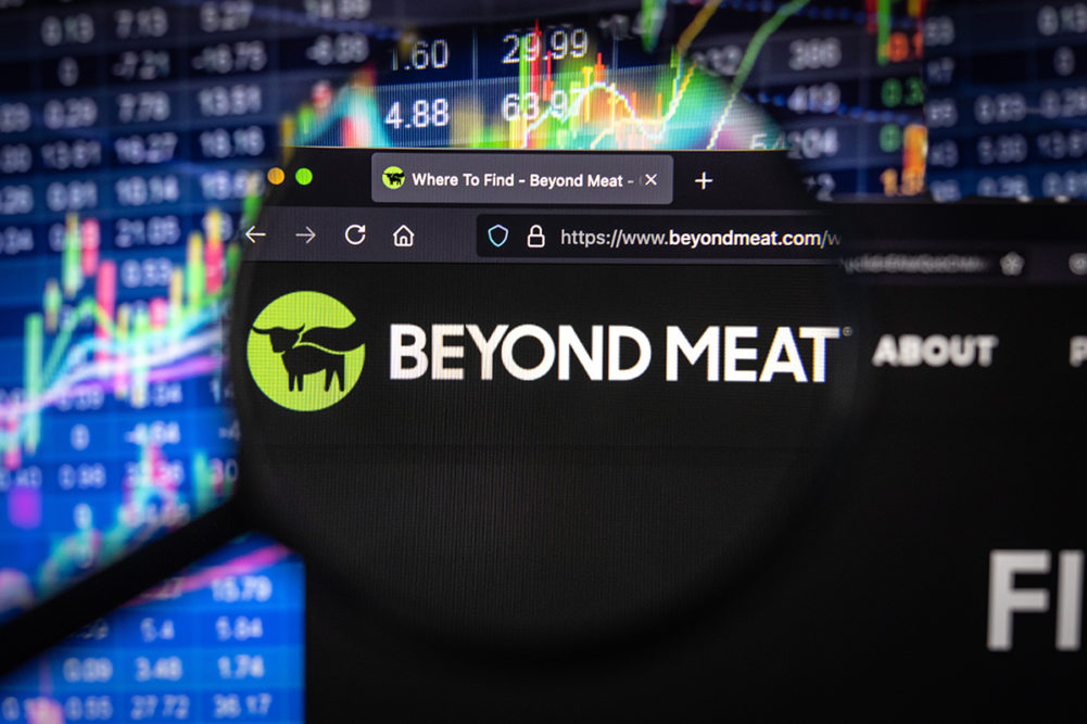 Beyond Meat company logo on a website with blurry stock market developments in the background, seen on a computer screen through a magnifying glass