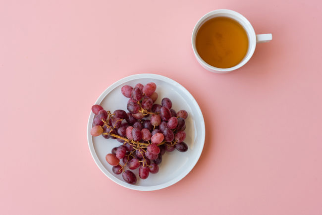 Directly above view of red grapes and white teacup on pink background