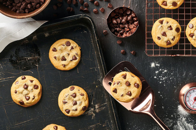 chocolate chip cookies on a counter with a bowl of chocoalte chips, a timer, and a spatula