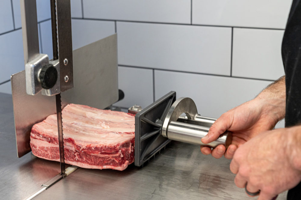 hands operating a meat saw with a piece of raw meat