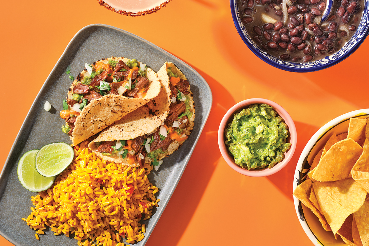soft tacos, rice, beans guacamole, and chips on a bright orange background