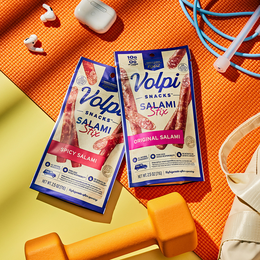 Volpi salami stix in packaging sitting on an orange yoga mat with an orange hand weight, wireless ear buds, blue jump rope, and tote bag on the mat around the product