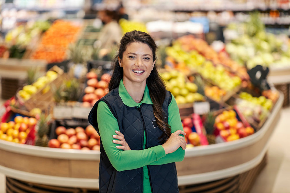 A smiling sales manager with arms crossed is standing at supermarket and smiling at the camera.