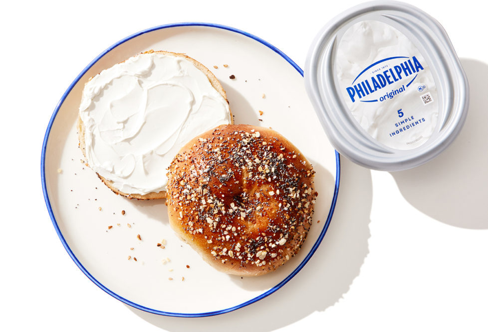 Philadelphia Cream Cheese container next to no-hole everything bagel covered in cream cheese