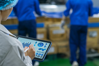 person holding a tablet with charts and graphs while people load shipping boxes in the background